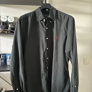 Svart skjorta från Ralph Lauren - Svart långärmad skjorta från Ralph Lauren i slim fit-modell. Skjortan har klassisk krage, vita knappar och den ikoniska röda broderade loggan på bröstet. Tillverkad i mjuk bomull som känns skön mot huden. Perfekt för dig som gillar stilrena och tidlösa plagg.