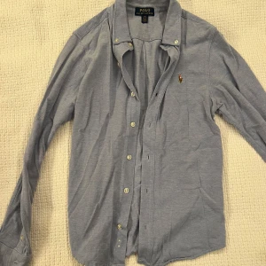 Ljusblå skjorta från Ralph Lauren - Snygg ljusblå skjorta från Ralph Lauren med klassisk krage och broderad polospelare på bröstet. Skjortan har långa ärmar, knappar framtill och är tillverkad i mjuk bomull. Perfekt för dig som gillar stilrena och tidlösa plagg. Bilden togs slarvigt men den är i väldigt fint skick 👍😊