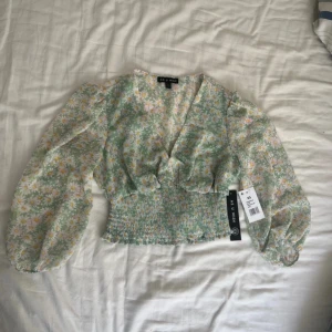 Blommig blus från As U Wish  - Supersöt blommig blus från As U Wish i ljusgrönt med vita och gula blommor. Blusen har v-ringning, långa puffärmar och smockad midja som ger en snygg croppad look. Perfekt för vår och sommar när du vill ha en romantisk vibe.
