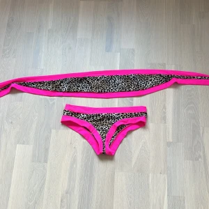 Leopardmönstrad bikini med neonrosa detaljer - Snygg bikini med leopardmönster och starka neonrosa kanter. Toppen knyter man själv som en tube top ungefär, bikinitrosan har samma coola leopardprint med rosa detaljer. Har aldrig använts! Skriv om ni bara vill ha trosan samma me toppen så sänks priset såklart. 