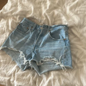 Levi's ljusblå jeansshorts med slitningar - Ljusblå jeansshorts från Levi's med råa, fransiga kanter och slitna detaljer framtill. Klassisk femficksmodell med knapp och dragkedja. Perfekta för sommaren och ger en avslappnad vibe. Levi's läderpatch baktill och röd etikett på fickan.
