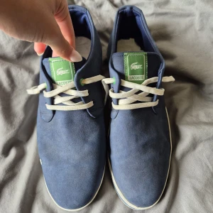 Blå sneakers från Lacoste - Snygga blå sneakers från Lacoste med vita snören och gröna detaljer. Skorna har en klassisk design med den ikoniska krokodilloggan på sidan. Perfekta för en stilren look.