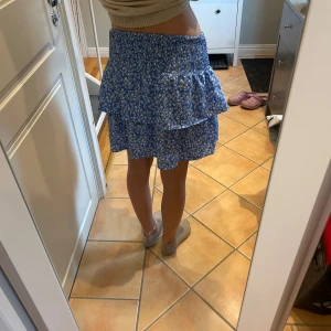 Blå blommig volangkjol - Supersöt blå kortkjol med vita blommor och bred smock i midjan. 