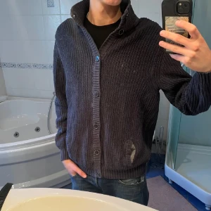 Mörkblå stickad kofta med krage - Säljer en mörkblå stickad kofta med grov ribbstickning och hög krage. Koftan har knäppning med stora knappar framtill och två fickor. Perfekt för lager-på-lager och chill stil. Passar dig som gillar en lite loose fit och klassisk look.