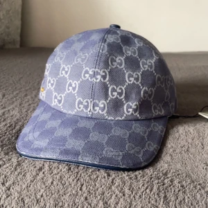 Gucci keps Dubai blå - Snygg blå keps från Gucci med ikoniskt GG-mönster över hela kepsen. Justerbar rem baktill i blått och klassisk böjd skärm. Tillverkad i bomullsmaterial med vit insida. Perfekt för dig som vill sticka ut med en lyxig accessoar.