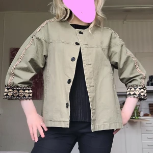 Jacka i khaki med broderade detaljer - Snygg khakigrön overshirt med broderade detaljer längs ärmarna och mönstrade muddar i svart och guld. Jackan har trekvartsärmar, knäppning framtill och en loose passform. Perfekt för dig som vill ha en unik och chill look. Passar xs-m men är L på lappen 🩷 knappt använd!