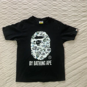 Svart BAPE t-shirt med camo-tryck - Svart t-shirt från BAPE med stort ikoniskt apehuvud i grått, blått och vitt camo-tryck på bröstet. Klassisk passform och korta ärmar. Tryckt text 'BY BATHING APE' under motivet. Perfekt för streetwear-stilen.