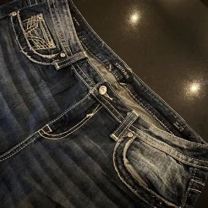 Vigoss bootcut jeans - Snygga blå jeans från Vigoss med bootcut-modell och kontrastsömmar. Bakfickorna har paljettdetaljer. Midjemått (rakt över midjan) - 48 cm, stussmått (rakt över stussen) - 58 cm, innerbenslängd - 82 cm💕