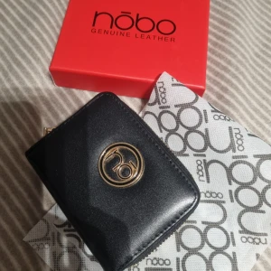 Svart skinnplånbok från Nobo - Stilren svart plånbok från Nobo i äkta skinn med guldfärgad logga framtill. Plånboken har dragkedja runtom, flera kortfack och tryckknappsstängning. Perfekt storlek för att få plats med det viktigaste och ändå ha en snygg accessoar i väskan.