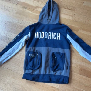 Blå och grå hoodie från Hoodrich - Snygg hoodie från Hoodrich i blått och grått med stora vita logotexter fram och bak. Tröjan har huva, långa ärmar med vita detaljer och två stora fickor med dragkedja framtill. Perfekt för en streetwear-look.