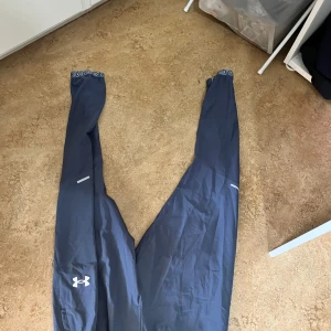 Blå träningsbyxor från Under Armour - Storlek S. Säljer ett par mörkblå träningsbyxor från Under Armour med elastisk midja och snörning. Byxorna har logga på benet och reflekterande detaljer. Materialet är syntet och passar perfekt för sport och träning. Snyggt tryck på midjebandet för extra stil.