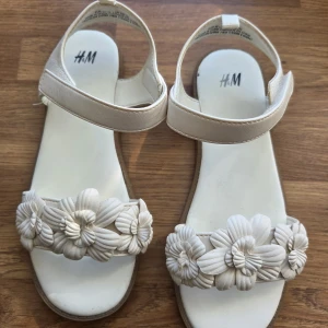 Vita sandaler med blommor från H&M - Snygga vita sandaler från H&M i storlek 32 med dekorativa blommor framtill. Sandalerna har en rund tå, platt sula och praktisk kardborreknäppning runt ankeln. Perfekta för varma dagar och ger en fräsch och somrig vibe till din outfit. Knappt använda, fint skick. 