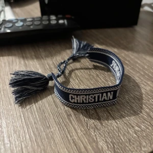Blå Christian Dior J'Adior armband - Snyggt blått armband från Christian Dior med texten 'CHRISTIAN DIOR J'ADIOR' broderad i vitt. Armbandet har dekorativa tofsar och knytning för justerbar passform. Perfekt accessoar för att lyfta din outfit med lite lyxig vibe.