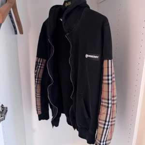 Svart Burberry hoodie med rutiga ärmar - Svart hoodie från Burberry i storlek M med klassiska rutiga paneler på ärmarna och logopatch på bröstet. Tröjan har huva, dragkedja och två fickor framtill. Perfekt för dig som vill ha en streetstyle med lyxig touch. Zipen är sönder!!!