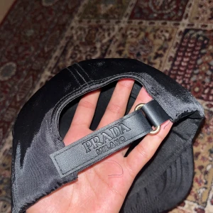 Svart keps från Prada Milano - Snygg svart keps från Prada Milano med glansig finish och ikonisk metalltriangel framtill. Justerbar rem med Prada-logga bak. Tillverkad i bomull och har en stilren, klassisk form som ger en lyxig vibe till din outfit.