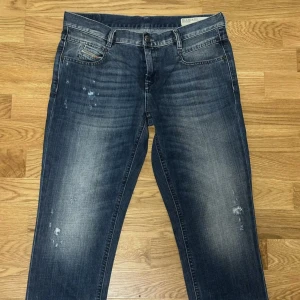 Diesel jeans blå W28 L32 - Snygga blå jeans från Diesel med slitningar och tvättade detaljer. Klassisk femficksmodell med dragkedja och knapp. Jeansen har en normal passform och är tillverkade i tåligt denim. Perfekta för dig som gillar en avslappnad och cool stil.