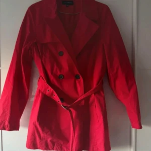 Röd trenchcoat från KappAhl - Snygg röd trenchcoat från KappAhl med klassisk krage, dubbelknäppning och bälte i midjan. Jackan har långa ärmar och svarta knappar framtill och baktill. Materialet är 100% bomull med foder i  polyester, perfekt för vår och höst. Stilsäker och tidlös modell. Använd en gång. Dubbelt storlek 40/42