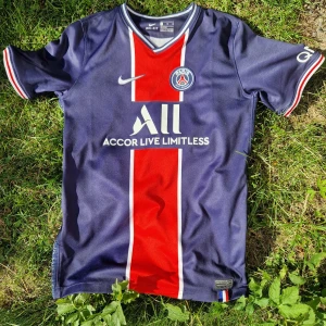 PSG Messi 30 Nike Dri-FIT XL tröja - Paris Saint-Germain fotbollströja med Messi 30 på ryggen. Mörkblå med röd och vit rand längs mitten, PSG-logga och Nike Swoosh på bröstet. Kortärmad, tillverkad i Dri-FIT material för bästa komfort. Snygga detaljer med klubbens 50-årsjubileum på sidan.