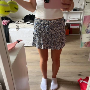 Blommig kortkjol från Zara XS - Supersöt kortkjol från Zara i storlek XS med blommigt mönster i blått, svart och vitt. Kjolen har volangdetaljer och en smockad midja med snörning framtill. Perfekt till vår eller sommar 💗säljer pga att den är för liten 