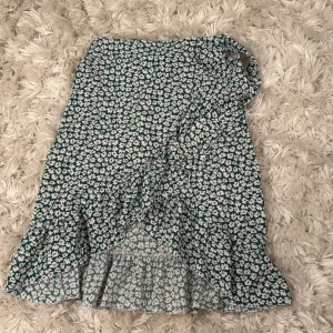 Grön blommig omlottkjol från SHEIN - Säljer en grön omlottkjol från SHEIN med vitt blommigt mönster och volangkant. Kjolen har knyt i midjan, en dragkedja och en snygg asymmetrisk form som ger en cool vibe. Perfekt för dig som gillar blommigt och vill ha något som sticker ut lite extra.