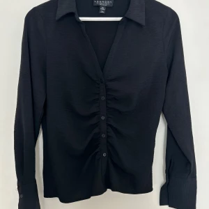 Svart skjorta  - Svart skjorta/ blus med rynkig i detalj i midjan vilket ger en superfin siluette. I mycket bra skick, säljer då den tyvärr inte kommer till användning. 