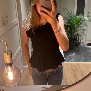 Svart blus med volang från JDY - Supersöt svart blus från JDY med broderade detaljer och volang nedtill. Ärmlös modell med rund hals och lätt croppad passform. Materialet är bomull och har en snygg struktur med små hål och mönster. 