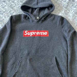 Supreme mörkgrå hoodie med logga - Supreme hoodie i mörkgrått med klassisk röd boxlogo på bröstet. Tröjan har huva, känguruficka och långa ärmar. Mjuk och skön bomullsmix som passar perfekt för chill dagar. Snygg streetwear-vibe och enkel att matcha med jeans eller joggers.
