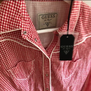 Rutig röd skjorta från Guess - Snygg oanvänd (prislapp kvar) röd och vit rutig skjorta från Guess med vita kontrastdetaljer och metallknappar. Skjortan har två bröstfickor och ett metallmärke med Guess-logga. 
