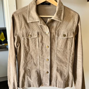 Beige manchesterjacka från Jasmin - Snygg beige manchesterjacka från Jasmin med klassisk krage och raka linjer. Jackan har långa ärmar, två bröstfickor med knappar och knäppning framtill med metallknappar. Perfekt för dig som gillar retrovibbar och vill ha en cool look.