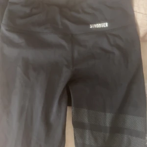 Svarta leggings från Stronger XS - Säljer ett par svarta leggings från Stronger i storlek XS. De har bred midja och coola mönstrade detaljer vid benen. Materialet är stretchigt med 75% återvunnen polyester och 25% elastan, perfekt för träning eller chill. Snygg logga framtill.