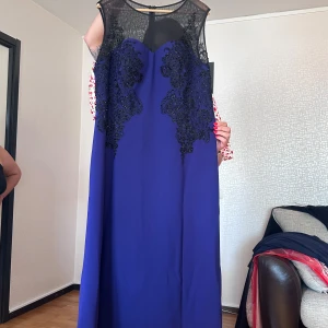 Lång blå klänning med spetsdetaljer - Säljer en elegant långklänning i djupblå färg med svart mesh upptill och vackra svarta spetsapplikationer med pärlor över bröstet. Klänningen har ärmlös design och en rak, svepande siluett som ger en lyxig känsla. Perfekt för bal eller fest