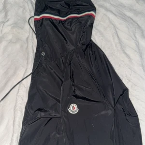 Svart vindjacka från Moncler - Svart vindjacka från Moncler med klassisk logga på armen och snygg randig detalj i rött, vitt och blått vid huvan. Jackan har dragsko i midjan och dragkedja framtill. Perfekt för dig som vill ha en stilren och sportig look.