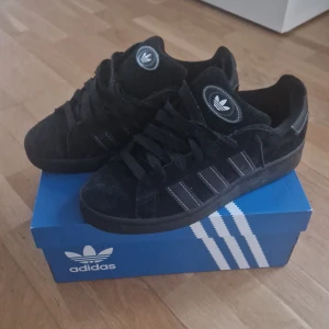 Svarta Adidas Campus sneakers mocka - Säljer ett par svarta Adidas Campus sneakers i mjuk mocka med klassiska tre ränder på sidan och Adidas-logga på plösen. Skorna har rund tå, platt sula och snörning. Perfekt för dig som gillar retrostil och vill ha något som funkar till streetwear.