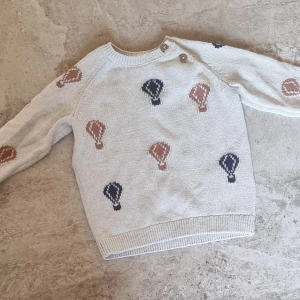 Stickad tröja med ballongmönster från Newbie - Superfin beige stickad tröja från Newbie med ballongmönster i marinblått och brunt, storlek 80.Tröjan har rund hals, två knappar vid axeln och ribbade muddar. Jag säljer ett par matchande leggings till i samma storlek.