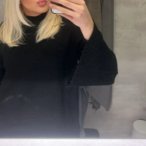 Svart stickad polotröja med vida ärmar - Säljer en svart stickad polotröja med breda, ribbade ärmar och lös passform. Tröjan har hög hals och är perfekt för dig som gillar en chill och oversized look. Materialet är mjukt och skönt, och den passar grymt till jeans eller leggings.