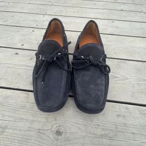 Mörkblå mocka loafers DelMare91 - Snygga loafers från DelMare91, tillverkade i Italien. Skorna är i mörkblå mocka med rund tå och har snörning framtill. Insidan är i ljusbrunt skinn och yttersulan är i svart gummi med coolt mönster. Perfekt för dig som gillar klassisk stil med en modern twist.