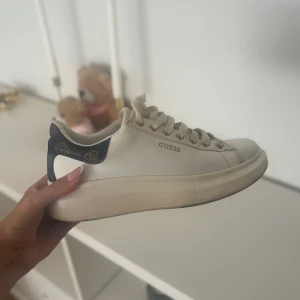 Guess vita sneakers med mönstrad häl - Snygga vita sneakers från Guess med rund tå och platt sula. Skorna har använts ett fåtal gånger o säljs då de aldrig kommer till användning. 