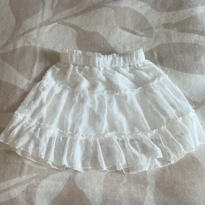 Vit volangkjol med prickigt mönster - Supersöt vit kortkjol med volanger och diskret prickigt mönster. Kjolen har resår i midjan och är gjord i ett lätt, luftigt material som ger en romantisk vibe. Perfekt för dig som gillar en feminin och trendig stil.