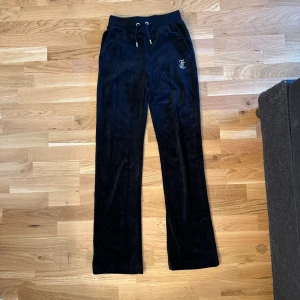 Svarta velourbyxor Juicy Couture XXS - Svarta velourbyxor från Juicy Couture i storlek XXS. Byxorna har snörning i midjan, raka ben och ikonisk Juicy-text med strass på baksidan. Perfekta för chill eller träning, med mjuk och glansig finish. Klassisk logga på framsidan och bekväm passform.