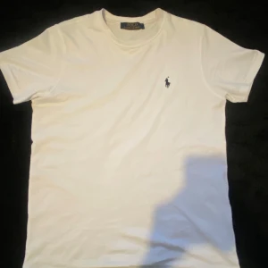 Vit t-shirt från Polo Ralph Lauren - Vit t-shirt från Polo Ralph Lauren med klassisk rund hals och korta ärmar. Diskret broderad logga i svart på bröstet. Mjuk och skön bomull som passar perfekt till jeans eller shorts. Enkel och stilren design som alltid funkar.