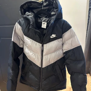 Svart och grå pufferjacka från Nike - Nike pufferjacka i svart och grått med coolt V-format mönster över bröst och rygg. Jackan har huva, dragkedja framtill och två sidofickor. Jackan är i storlek S och för barn men står L i jackan. 