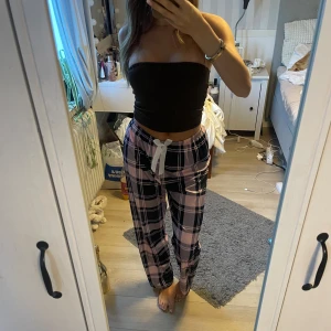 Rosa och svarta rutiga mjukisbyxor - Säljer ett par mjukisbyxor med rosa och svarta rutor, perfekt för chill dagar hemma. Byxorna har en vit snörning i midjan och är i loose fit med raka ben. Materialet känns mjukt och skönt mot huden. Passar dig som gillar avslappnad stil och snygga mönster. Aldrig använda