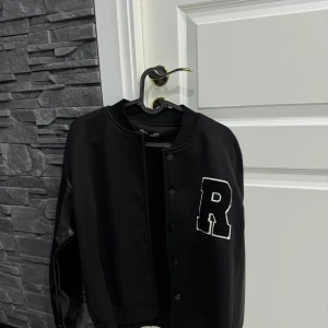 Svart bomberjacka med R från SHEIN - Svart bomberjacka från SHEIN med stora vita R-trycket på bröstet. Jackan har svarta knappar framtill och coola ärmar i skinnimitation. Ribbad krage och muddar ger en snygg streetstil. 