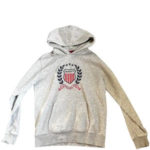 Grå hoodie från GANT med emblem - Snygg grå hoodie från GANT med stort broderat emblem på bröstet i blått och rött. Tröjan har huva, känguruficka och ribbade muddar. Perfekt för dig som gillar klassisk college-stil och vill ha något bekvämt och stilrent.