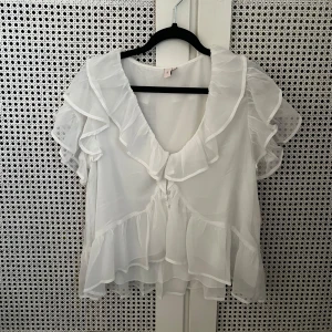 Vit volangblus från Nelly - Aldrig använd. Strl 36 Supersöt vit blus från Nelly med transparenta volanger längs halsringning, ärmar och nederkant. Blusen har v-ringad front och korta ärmar med extra volym. Perfekt för dig som gillar romantisk och feminin stil.