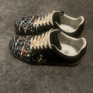 Maison Margiela  - Maison Margiela sneakers i svart skinn med coola färgstänk i blått, rött, gult, grönt och vitt över hela ovandelen. Skorna har beige snörning och en klassisk gummisula i brunt. Insidan är vit och designen är riktigt unik och konstnärlig.