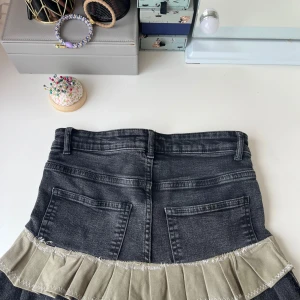 Svart jeanskjol med beige volang - Cool svart jeanskjol med två stora bakfickor och klassiska framfickor. Kjolen har en beige volangdetalj längst ner som ger en unik och edgy vibe. Passar perfekt till sneakers eller boots och har bälteshällor för extra stylingmöjligheter.