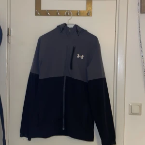 Under Armour grå och svart vindjacka - Snygg vindjacka från Under Armour i grått och svart med huva och dragkedja hela vägen. Jackan har en bröstficka med dragkedja och logga på framsidan. Perfekt för träning eller chill ute. Långärmad och med justerbar huva för extra komfort.