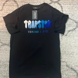 Trapstar svart t-shirt med blått tryck - Svart t-shirt från Trapstar med stort broderat Trapstar-tryck i vitt och olika blå nyanser på bröstet. Under trycket står det 'IT'S A SECRET' i blått och vitt. Klassisk rund hals och korta ärmar. Perfekt för dig som gillar streetwear och snygga detaljer.