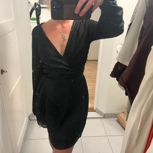 Svart omlottklänning med snörning - Snygg svart kort omlottklänning med långa ärmar och v-ringad hals. Klänningen har tunna vertikala ränder och ett knytband i midjan som ger en smickrande siluett. Materialet är lätt och har en diskret glans, perfekt för dig som gillar stilrena detaljer.